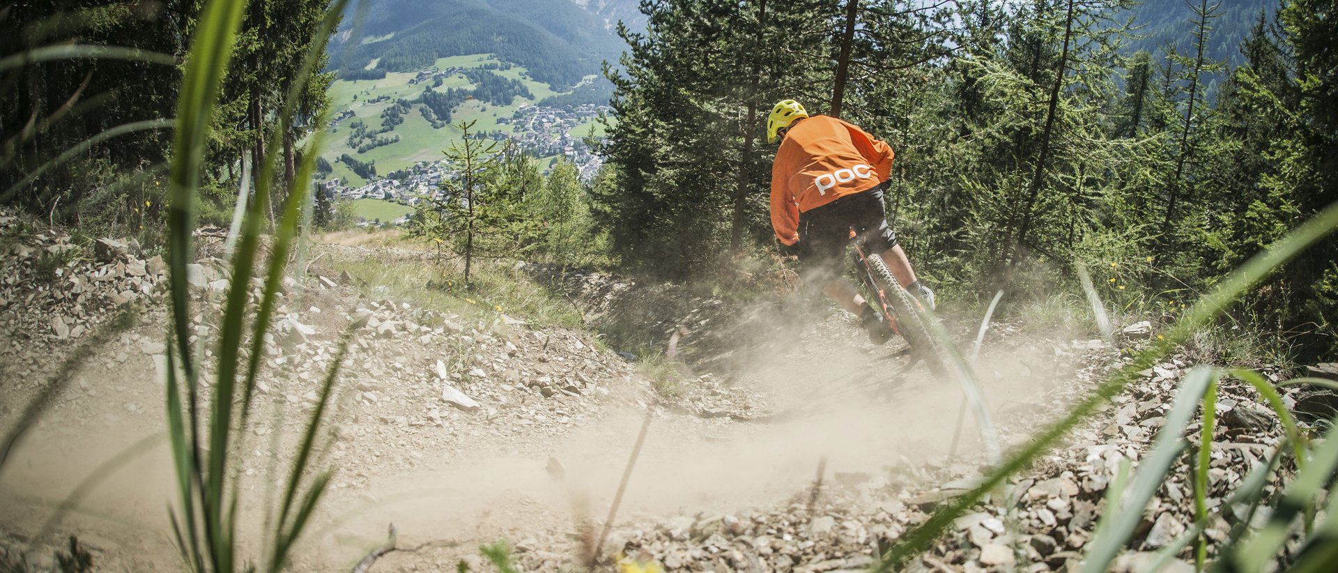 Active holidays in South Tyrol: a great experience Mountain biker rides dusty trail in forested mountain area with village in background
