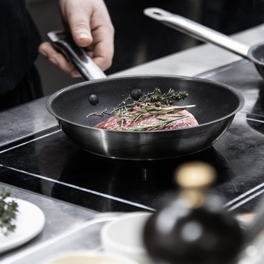 Unser familiengeführtes Hotel in Südtirol Hand hält Pfannengriff, Fleisch mit Kräutern in Pfanne auf Herd gebraten