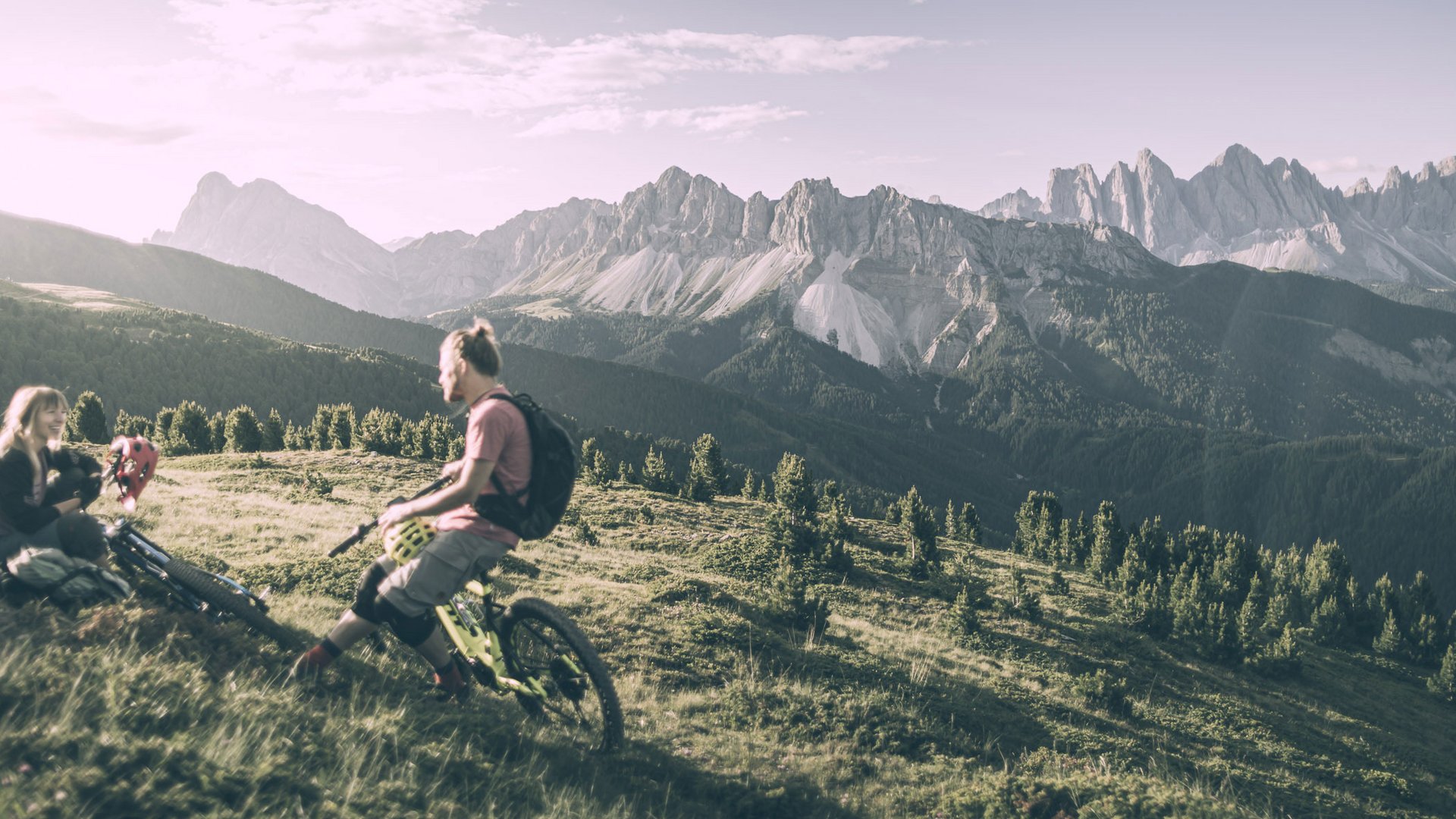 Biking at Plan de Corones/Kronplatz: unique Two mountain bikers resting on a meadow with mountain landscape in the background