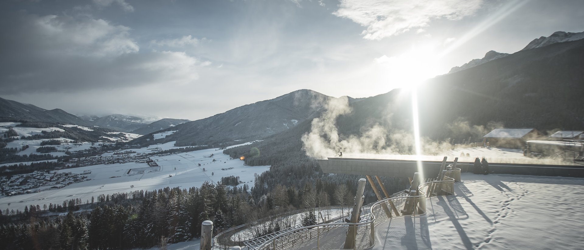 Our wellness hotel in Val Pusteria for relaxation seekers Sunny winter view of snow-covered mountains and valley with smoke above rooftop