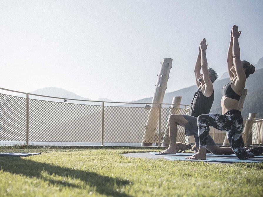 Our 5-star hotel in Italy between heaven and earth Two people practicing outdoor yoga on grass with a mountain view.