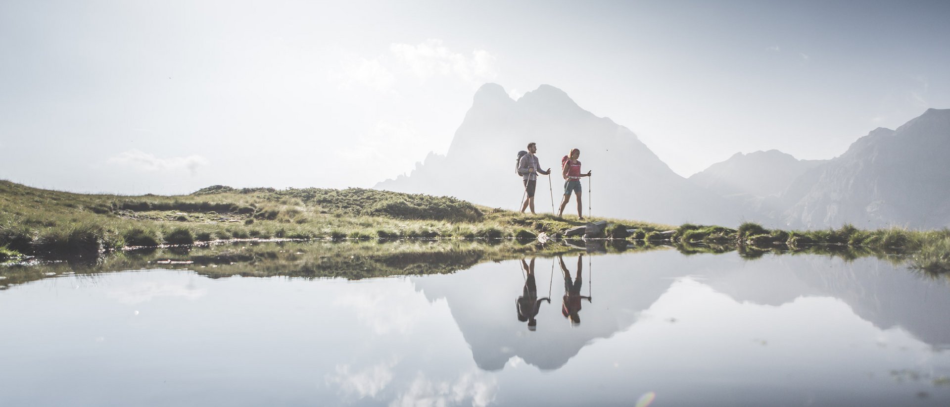 Our 5-star hotel in Italy between heaven and earth Two hikers with poles by a mountain lake with reflected mountains in the background