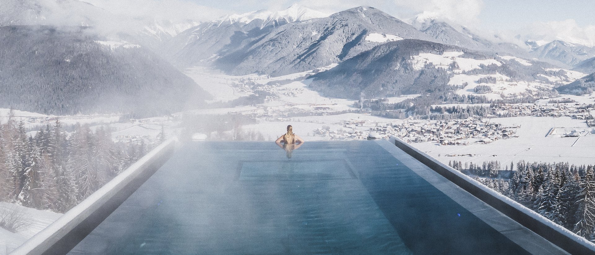 Unser aktueller Katalog zum Downloaden Person im beheizten Infinity-Pool mit Blick auf verschneite Berge und Tal