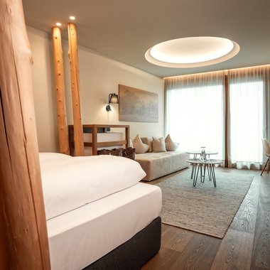 Your 5-star hotel in the Dolomites Modern hotel room with bed, sofa, tables, and large window with curtains